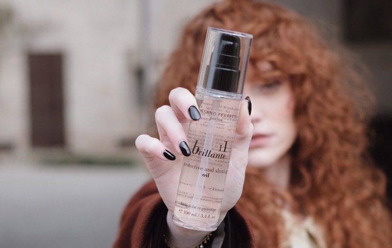 Image of a curly redhead girl showing Rossano Ferretti Parma's Brillante protect and shine oil in her hands.