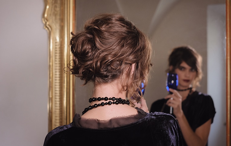 Image of a girl with an elegant chignon looking at herself in the mirror while holding a glass in her hand.