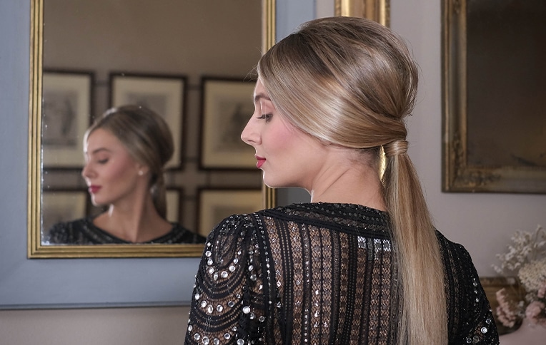 Image of a blonde girl with straight hair in a ponytail looking at herself in the mirror.
