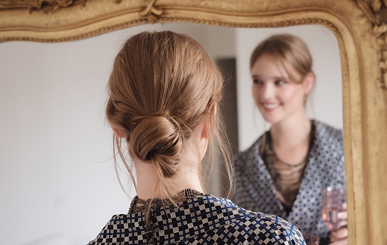 Image of a girl with an elegant chignon looking at herself in the mirror while holding a glass in her hand.