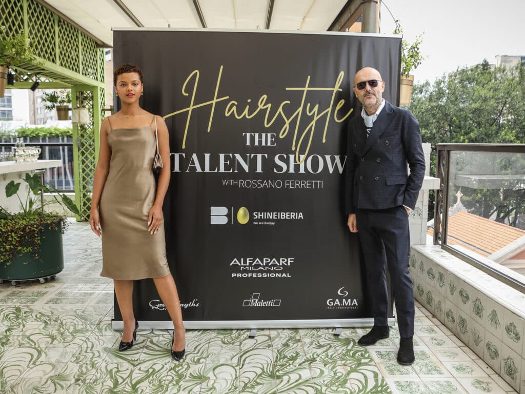 An image of Rossano Ferretti posing in front of a billboard for his TV show, "Hairstyle: The Talent Show," alongside an influencer.