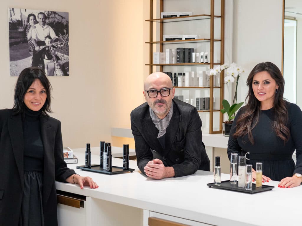 Image of Rossano Ferretti in one of his salons with two other women and various products from his brand Rossano Ferretti Parma.