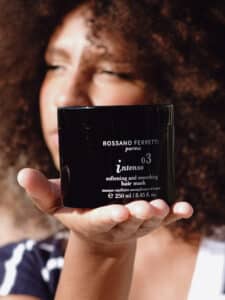 Image of a girl with brown, curly hair holding the Intenso smoothing mask by Rossano Ferretti Parma in her hand.