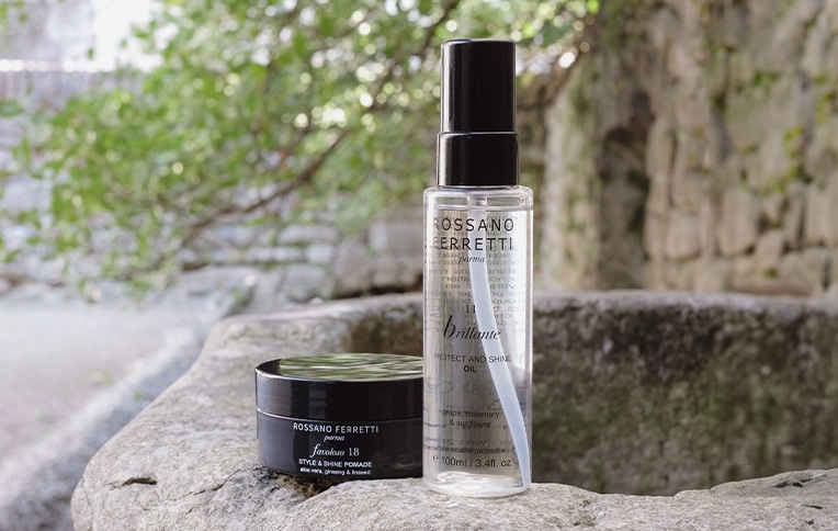 Image of Rossano Ferretti Parma products, featuring the Favoloso Style & Shine Pomade and Brillante Protect & Shine Oil. The products are placed on top of a small wall set against a backdrop of trees. 