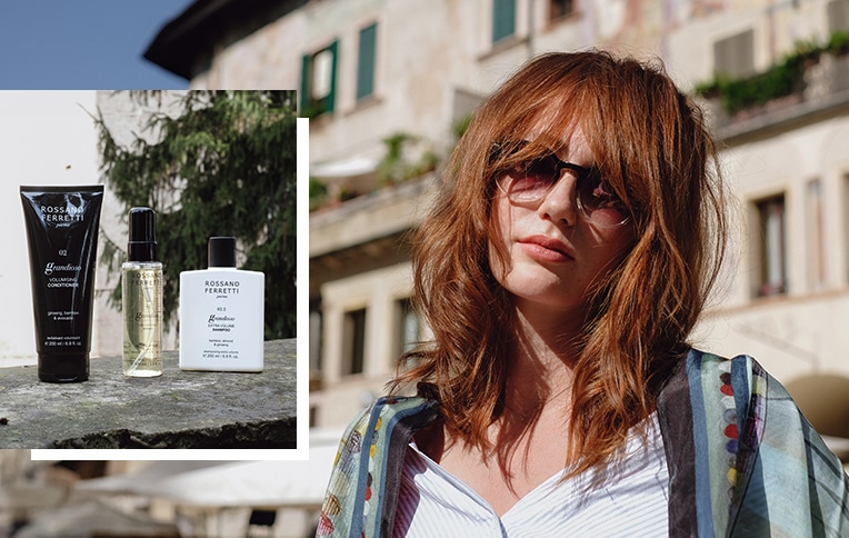 Ragazza rossa con capelli voluminosi accanto alla routine volumizzante Grandioso di Rossano Ferretti Parma, che include lo shampoo extra volume, il balsamo volumizzante e lo spray volumizzante.