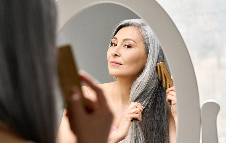 Woman with long gray hair combing her hair in front of a mirror.